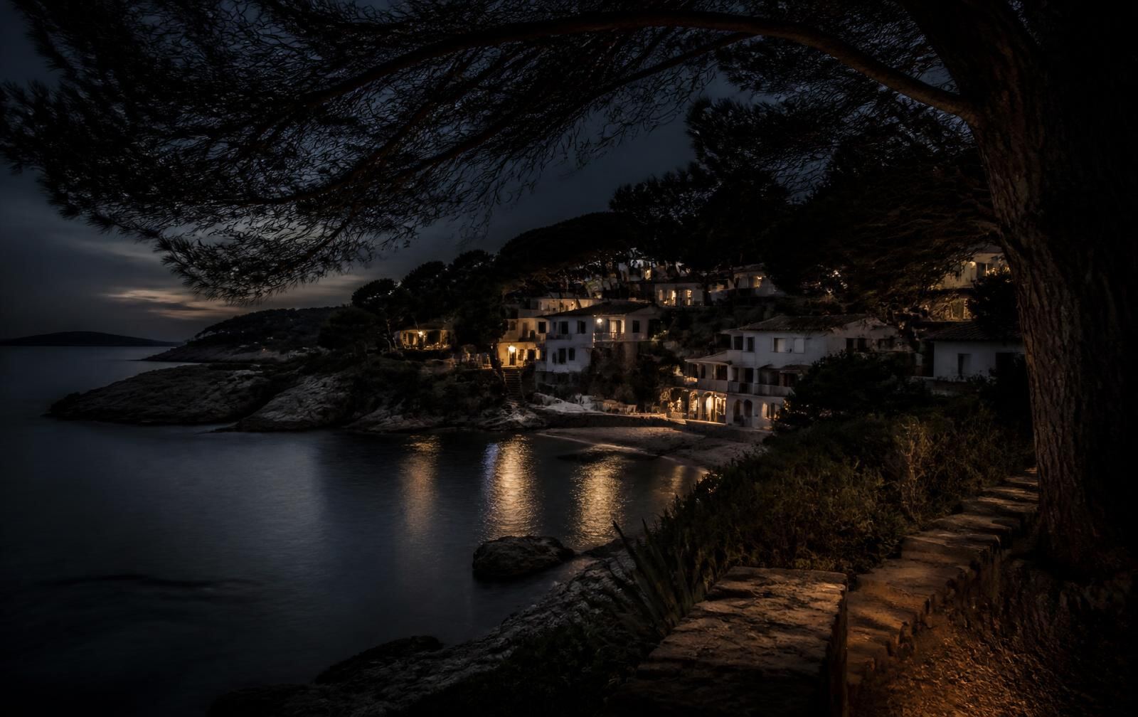 A pine-framed Costa Brava cove at twilight with a small whitewashed village lit warm gold above dark water