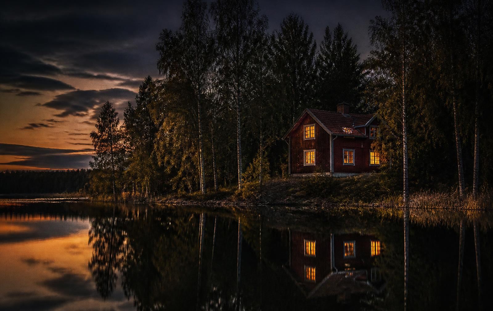 A Swedish countryside lake at late summer evening, a dark red timber house with warm gold windows among birches reflected in still water
