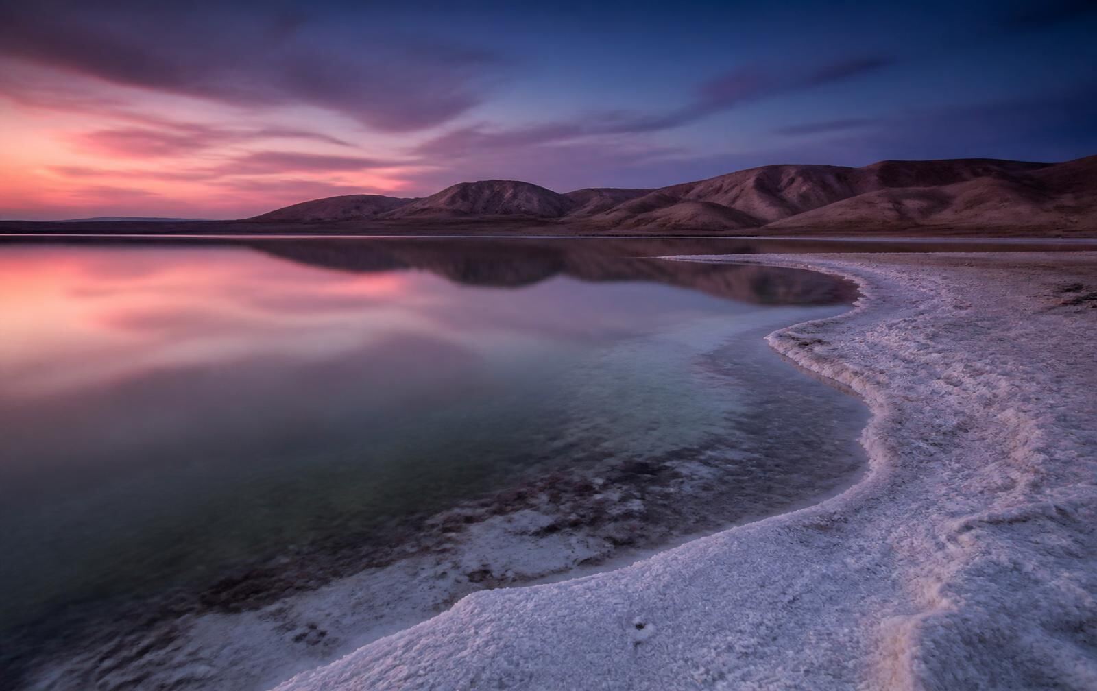 Still pale salt lake at dusk with a crusted white mineral shoreline and bare arid hills beyond