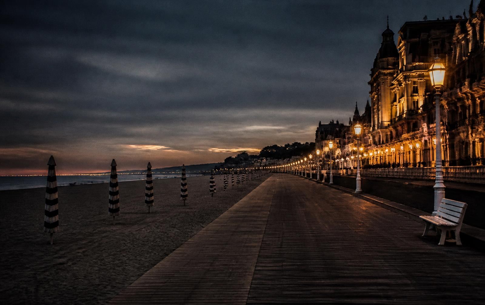 The Deauville seaside boardwalk at dusk with closed parasols, wide sand and lamplit belle époque facades