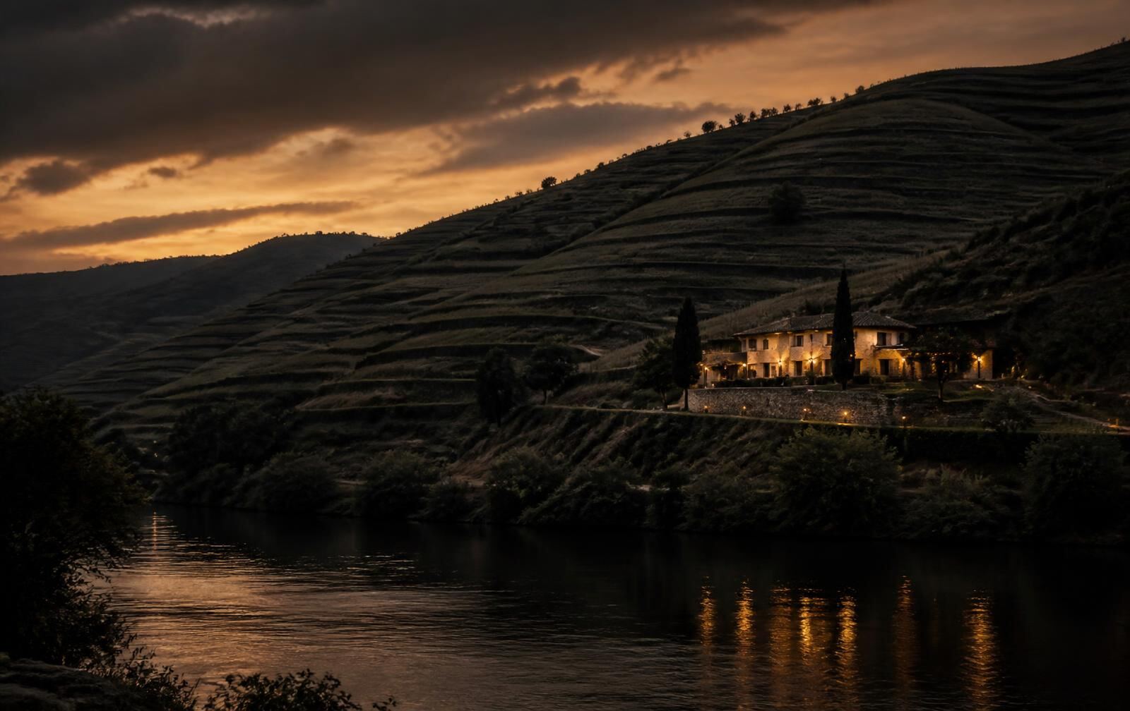 Terraced vineyard hillsides above a dark river at dusk with one lit house on the slope