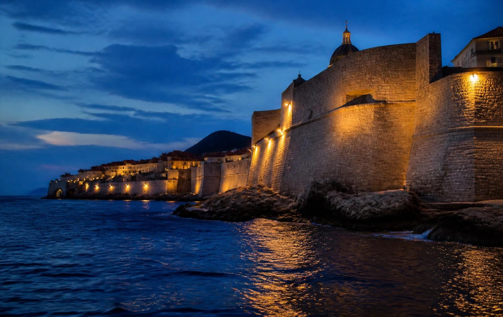 Dubrovnik's floodlit city walls rising from a dark Adriatic at dusk, warm lights along the ramparts
