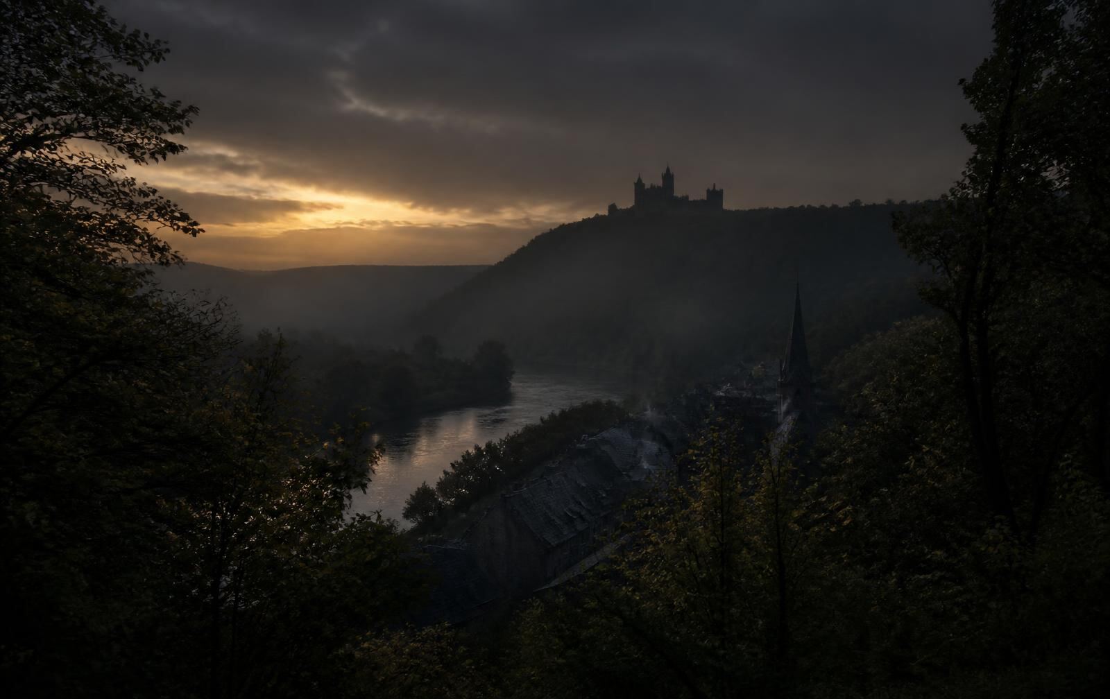 A wooded northern river valley at dusk with a distant castle silhouette on a ridge above a small town