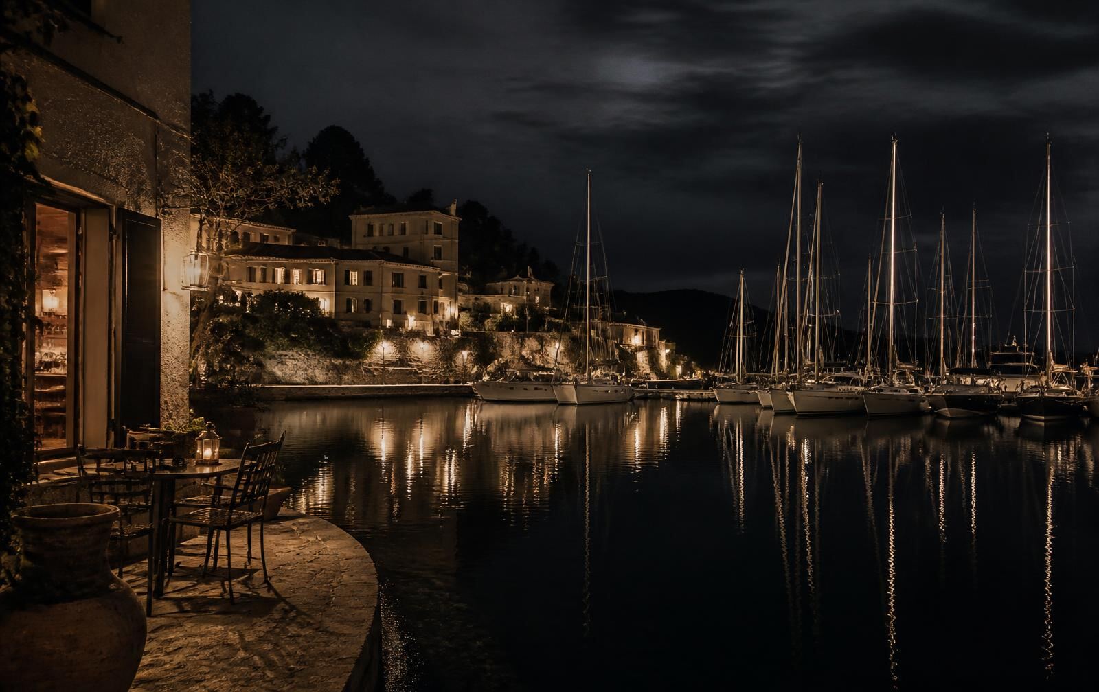 A quiet Mediterranean marina at night with mast lights reflected on black water and a warm coastal interior nearby