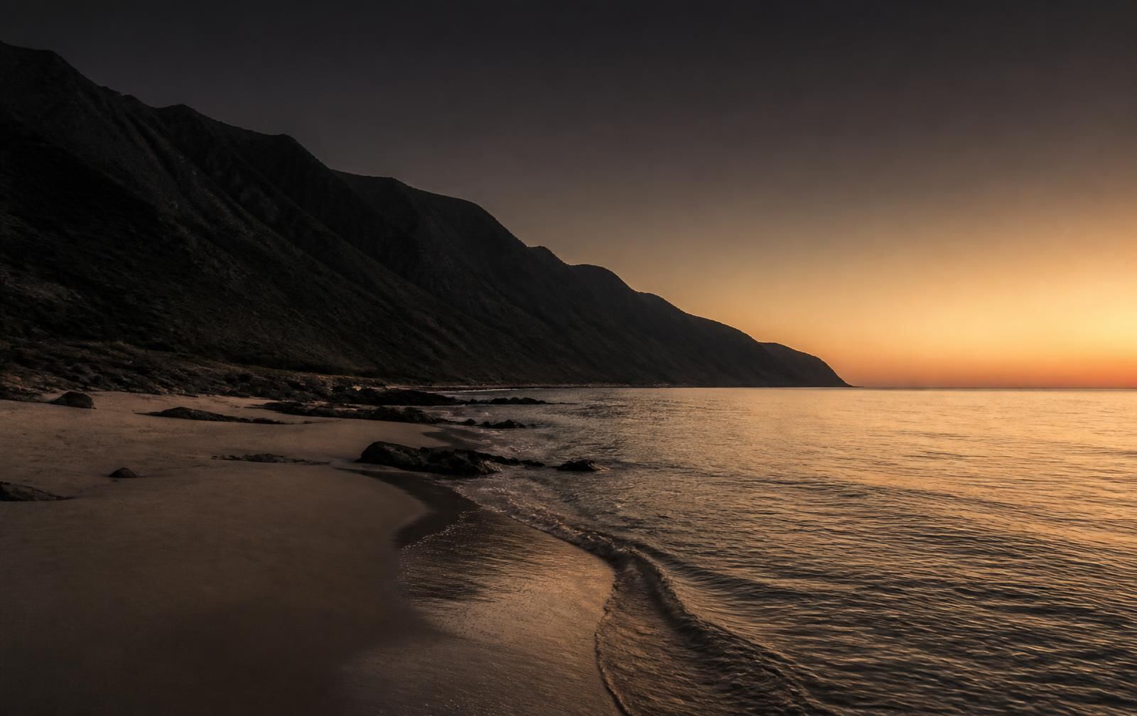 A rocky east-coast shoreline at dusk with dark mountains falling into a calm sea and warm gold horizon light