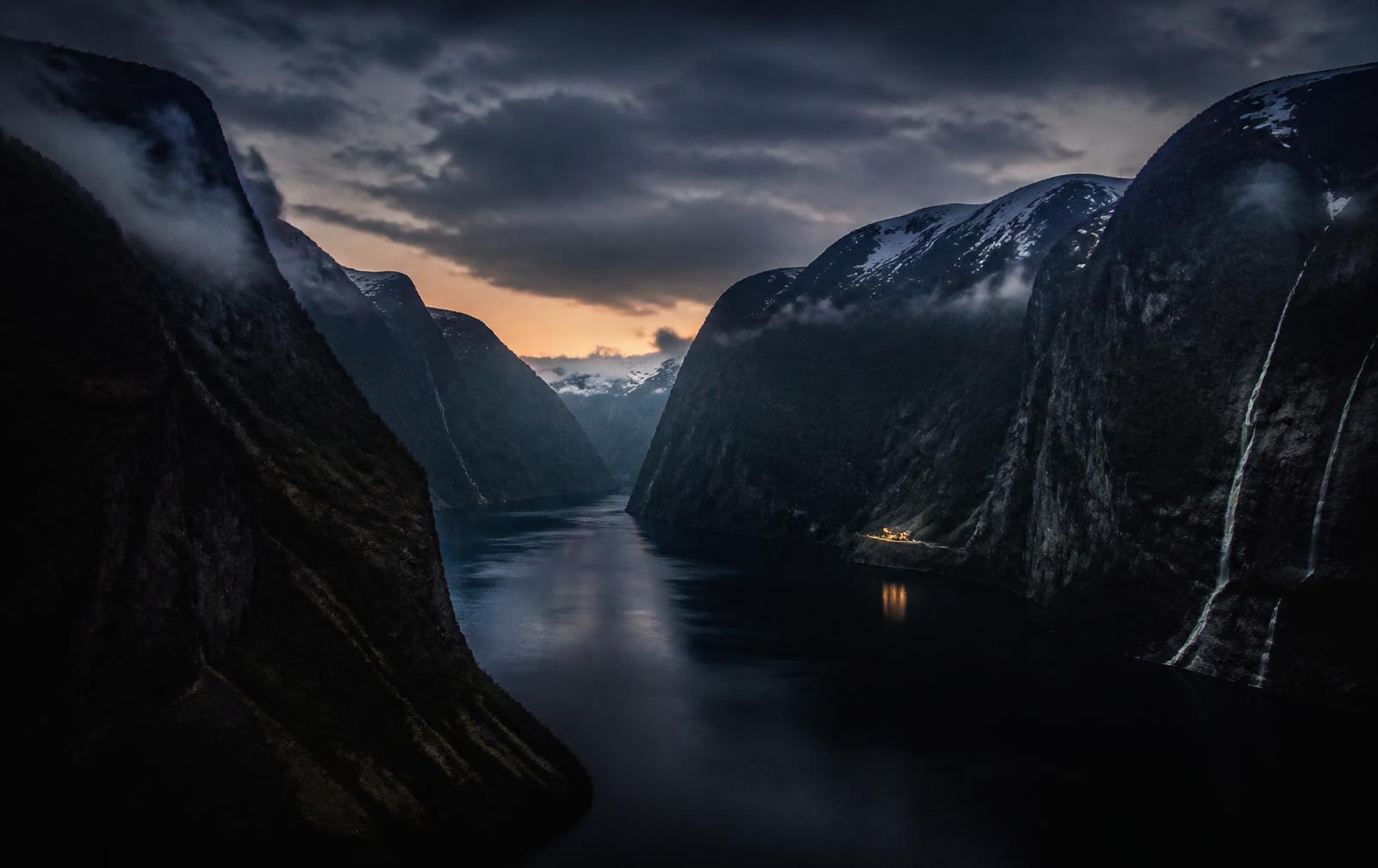 A deep narrow Norwegian fjord at dusk, sheer dark cliffs and thin waterfalls above black mirror water, one small warm-lit building far below