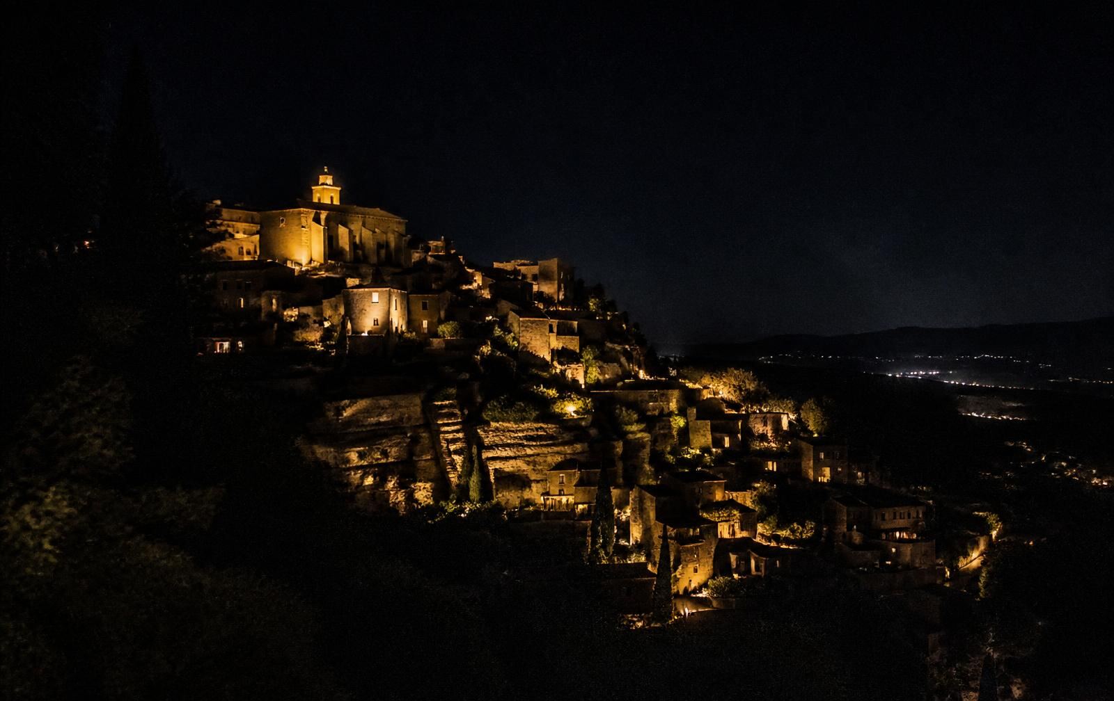The hilltop village of Gordes lit gold at night above a dark Luberon valley