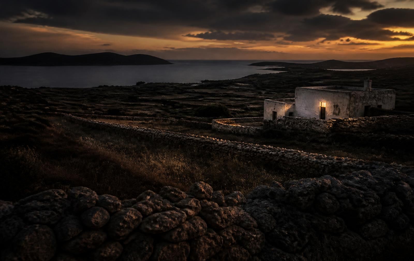 A quiet island landscape at dusk with dry stone walls, low countryside and a limestone farmhouse with one lit window near the sea