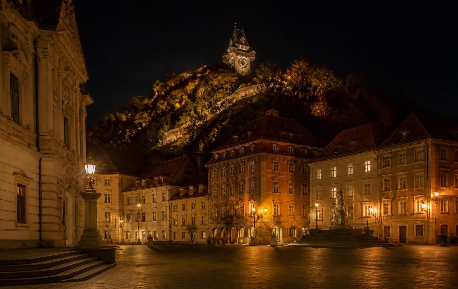 A historic Graz square at night with warm gold lamplight on stone and the clock tower hill above