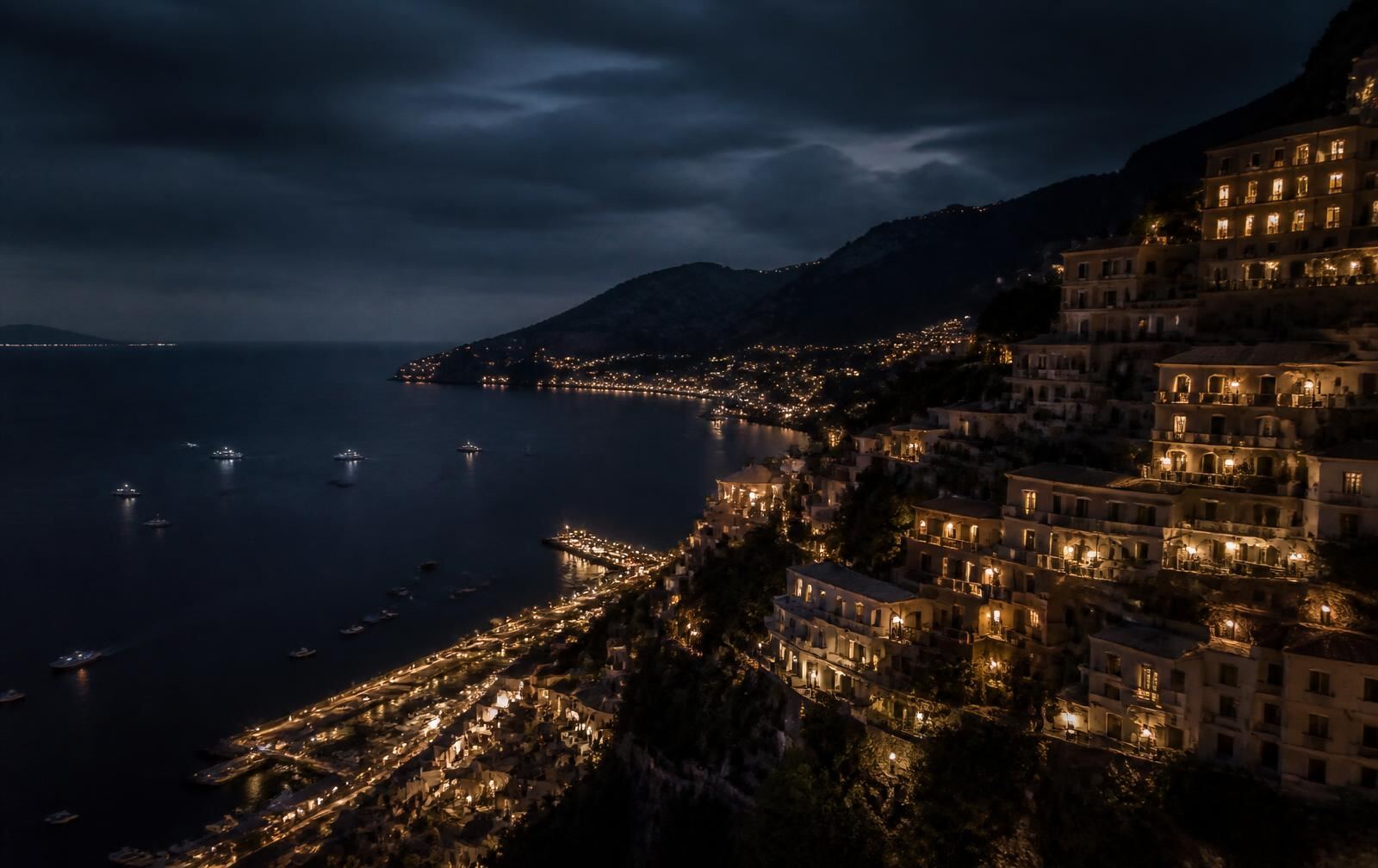 Hillside city at night descending in tiers of warm lights towards a dark Mediterranean bay