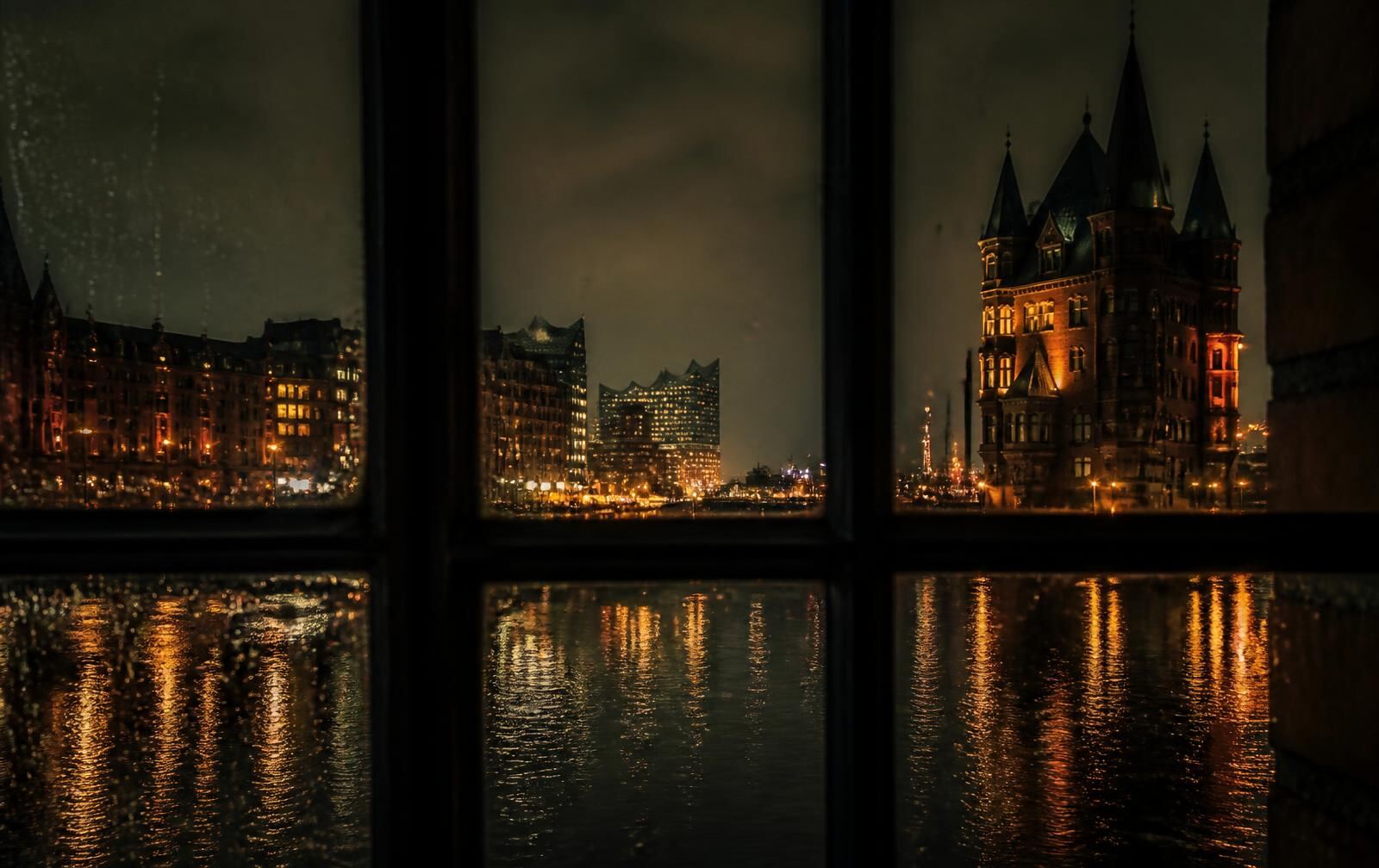 Hamburg at night in the rain, brick warehouse facades reflected in canal water with warm light behind tall windows