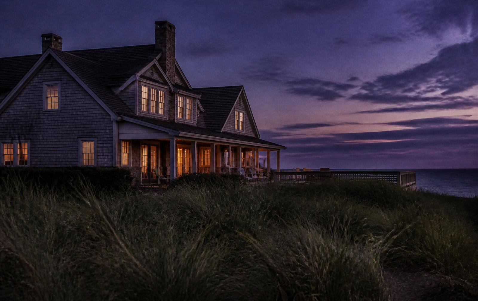 Shingled Hamptons house with warmly lit windows behind dune grass at summer dusk