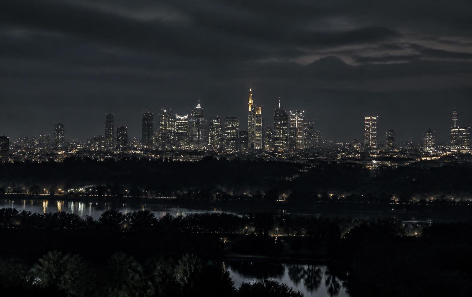 A distant illuminated European skyline across a dark river plain and wooded hills at night