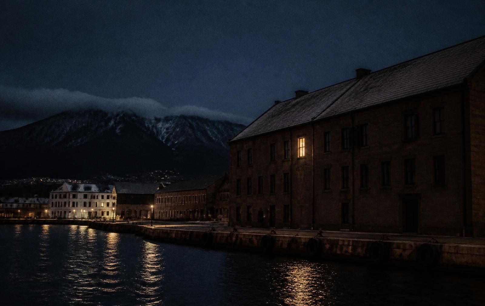 Hobart's waterfront at night with dark water, sandstone warehouses, a mountain silhouette and one warm lit window