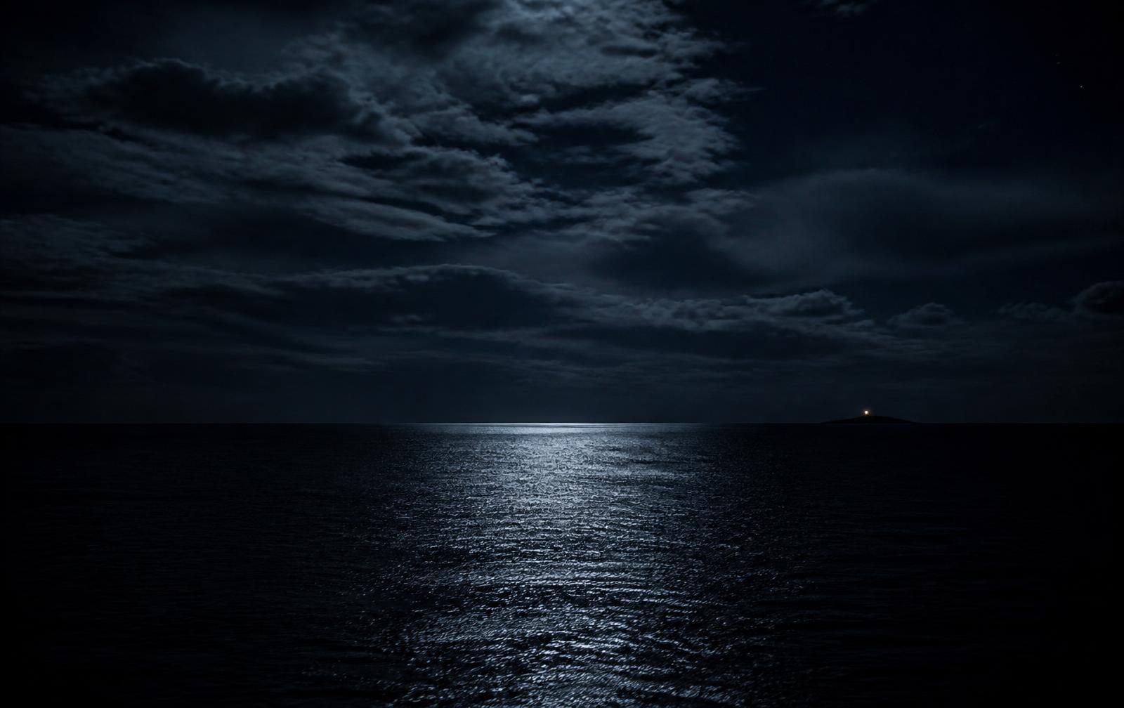 A vast dark ocean at night with a narrow band of moonlight and one distant island light