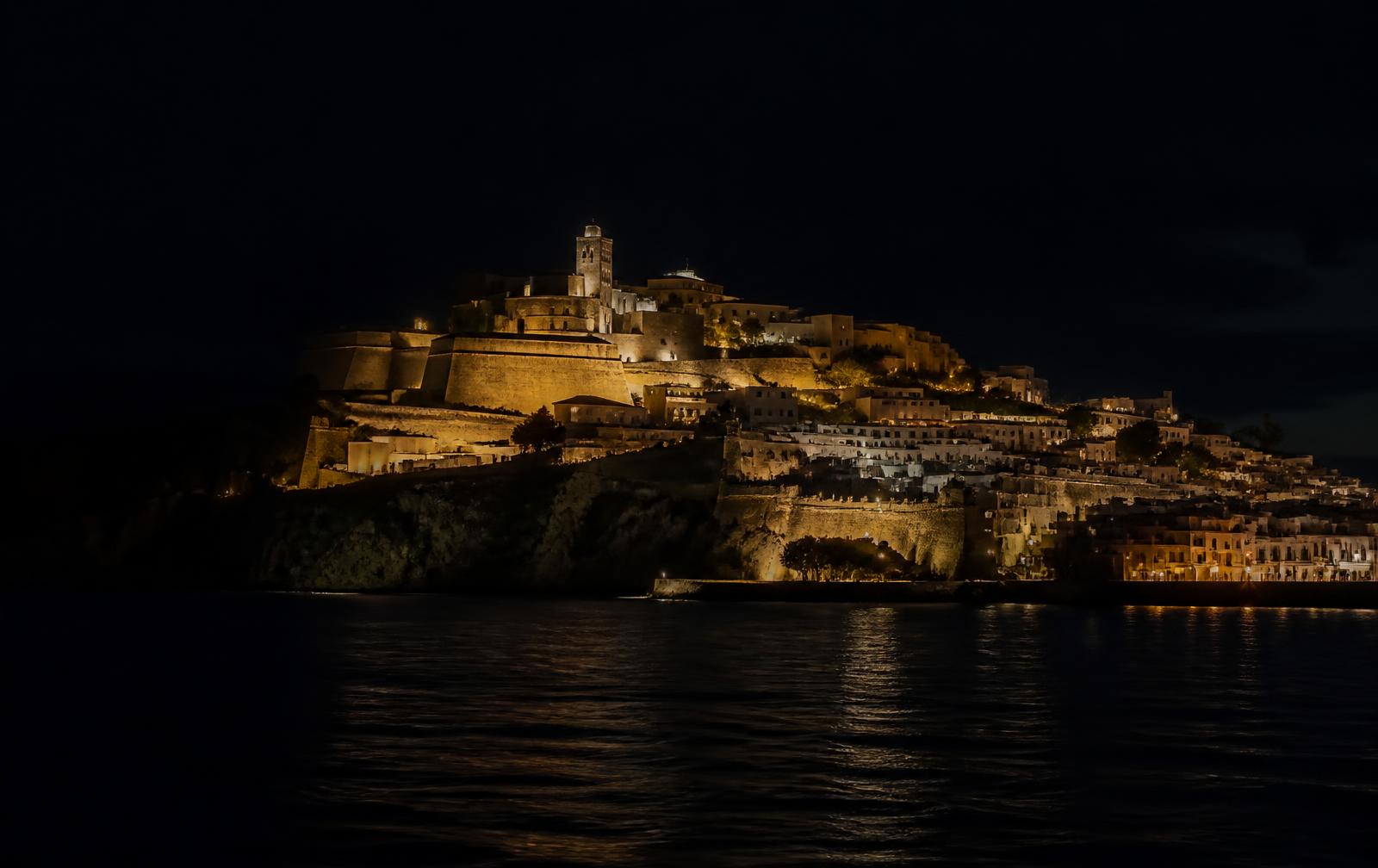 The fortified old town of Ibiza lit in warm gold at night above a dark Mediterranean bay