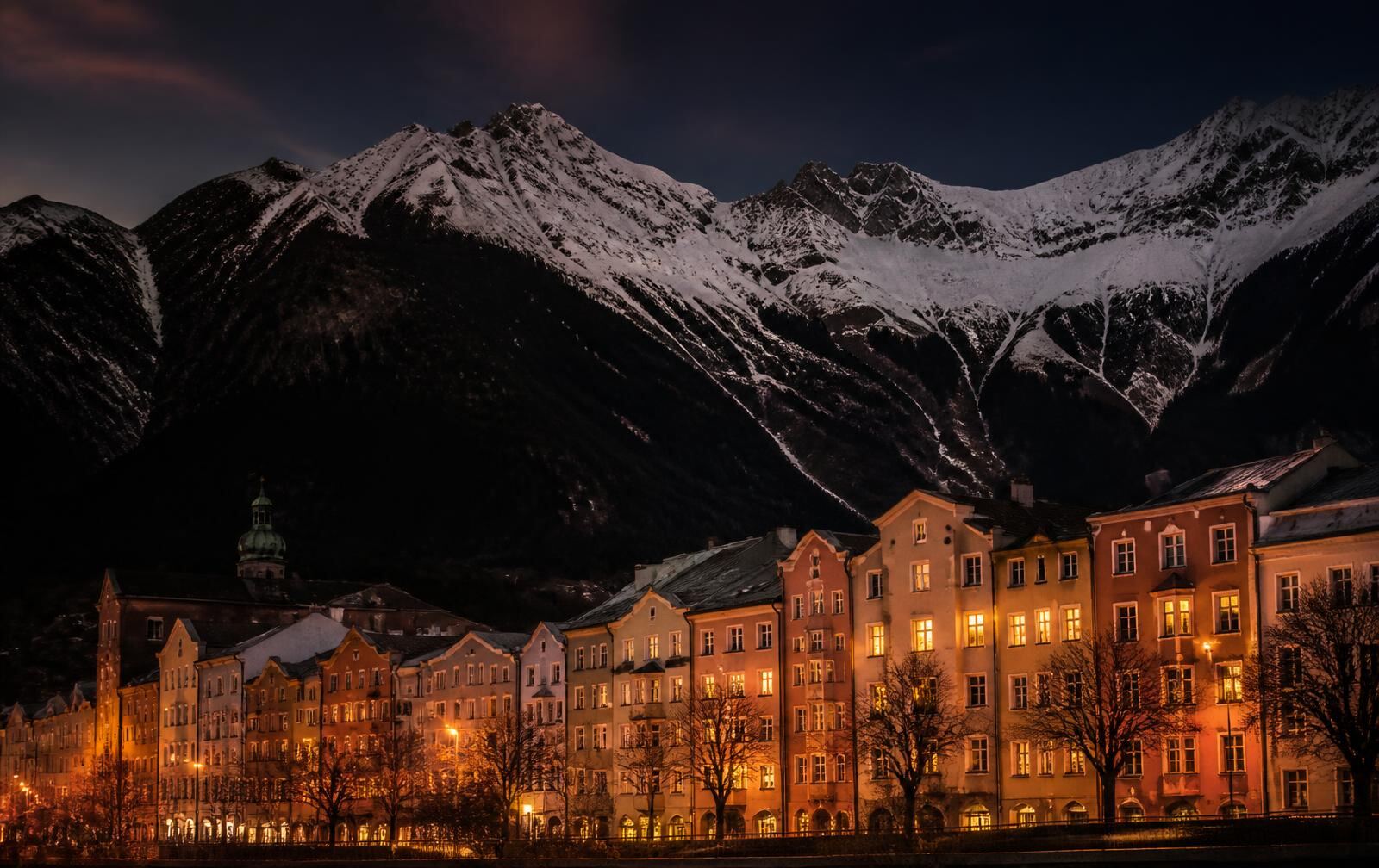 Innsbruck's lit riverside facades at dusk beneath the towering snow-covered Nordkette range