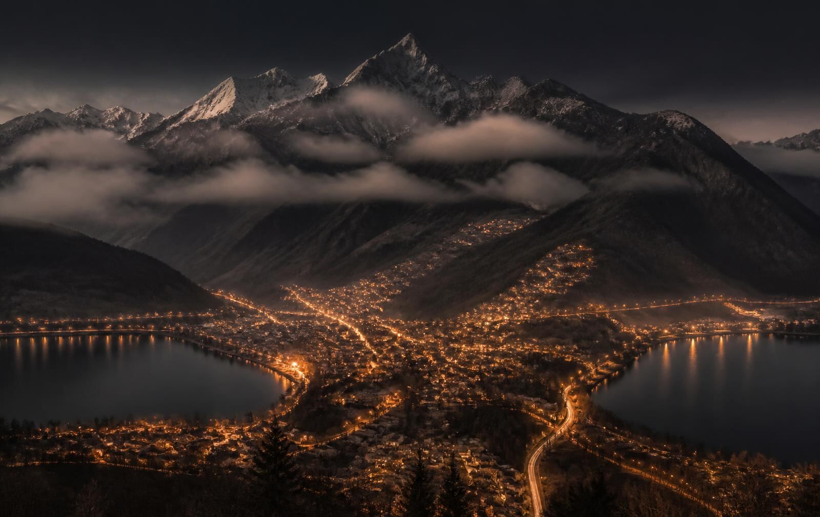 A valley town between two dark lakes at twilight with high snow peaks behind and warm gold lights below
