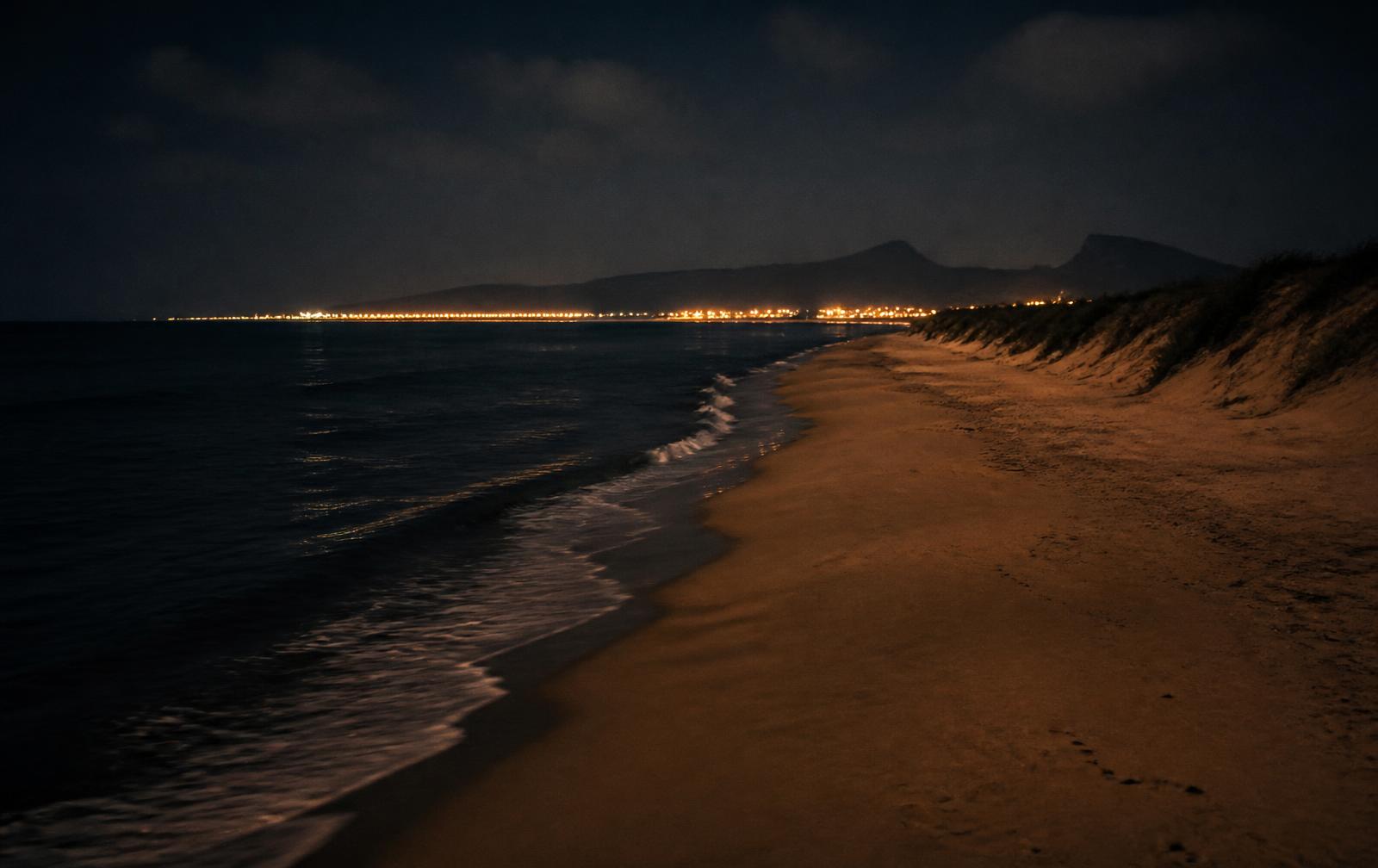 Long empty sandy Mediterranean beach at night with dark surf and distant coastal lights