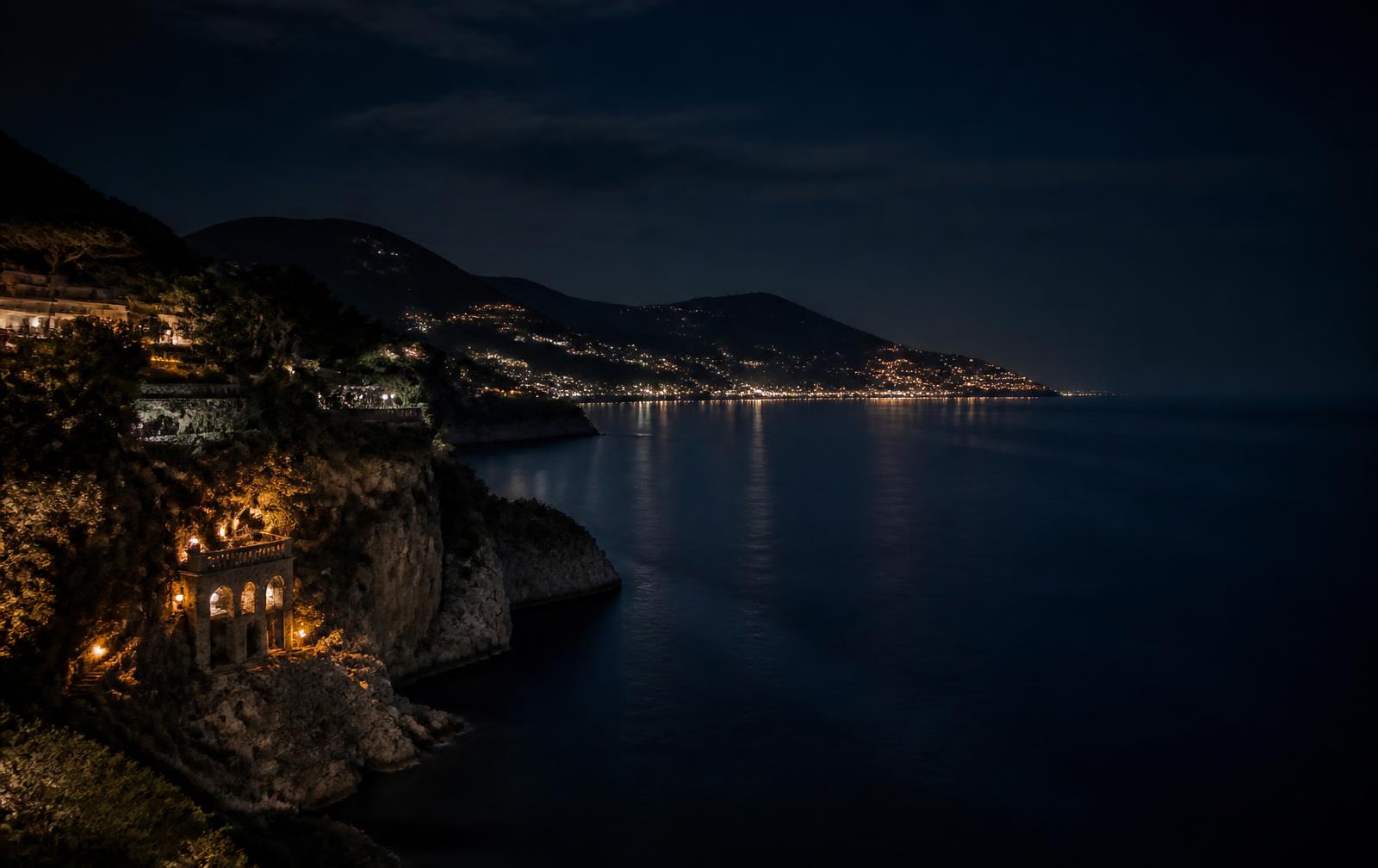 Mediterranean coastline at night with warm distant town lights reflecting across dark calm water