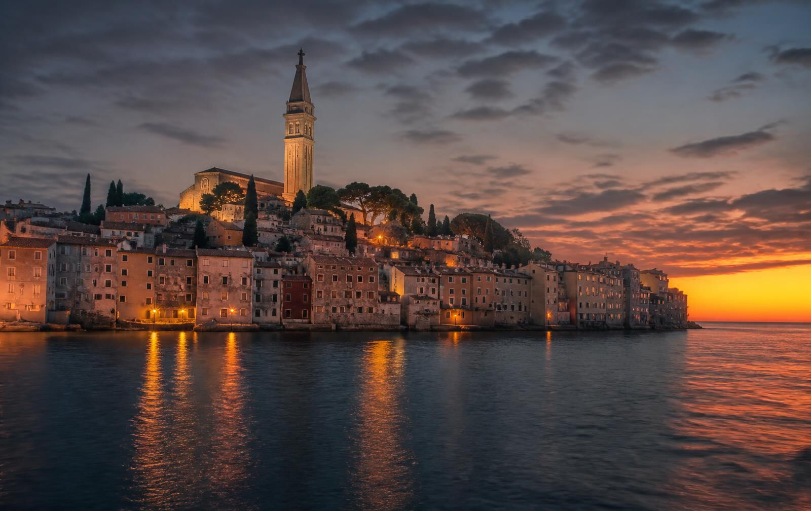 An Istrian hill town at dusk — stone tower and rooftops above green hills under a deep purple evening sky