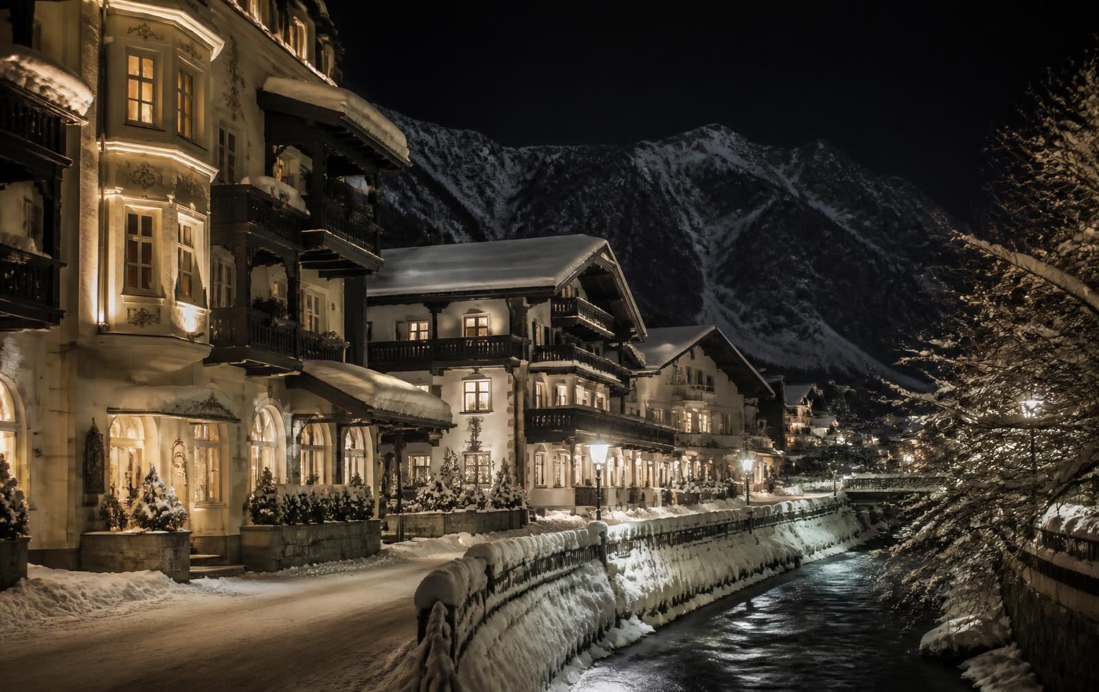 A snow-covered Tyrolean old town street at night with warm gold light spilling from windows below dark peaks