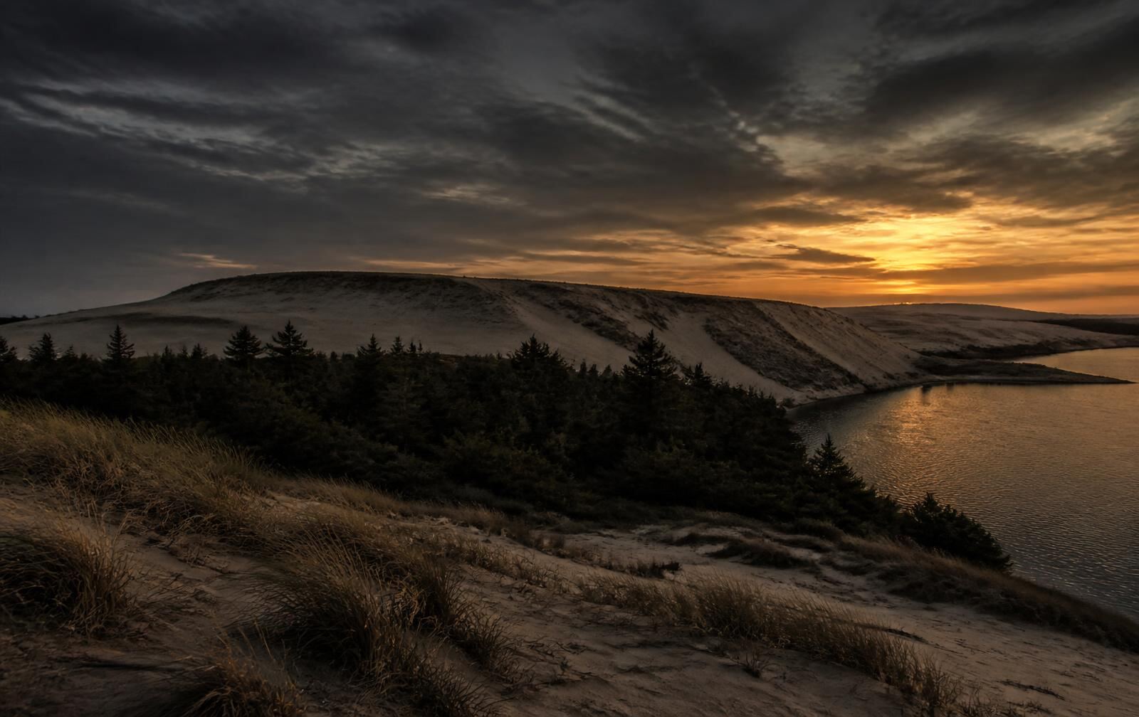 Curonian Spit at dusk, high pale sand dunes above dark pine forest with the still Curonian Lagoon below and a heavy grey and amber sky