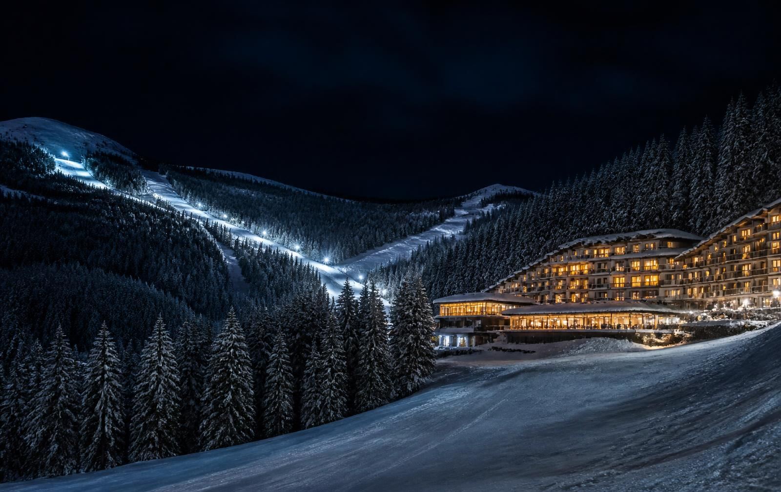 A snow-covered Balkan ski resort at night with floodlit pistes and a hotel glowing warm gold below dark pines