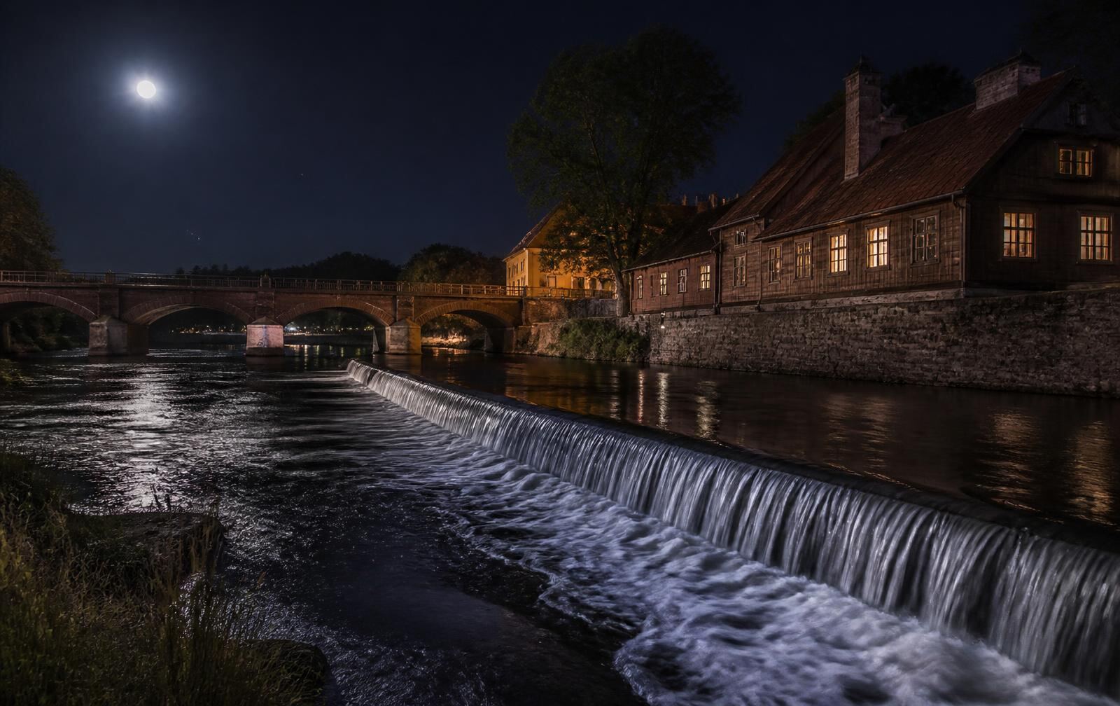 Kuldīga at night — the wide Venta waterfall glimmering under moonlight, an old brick bridge and warm-lit timber houses