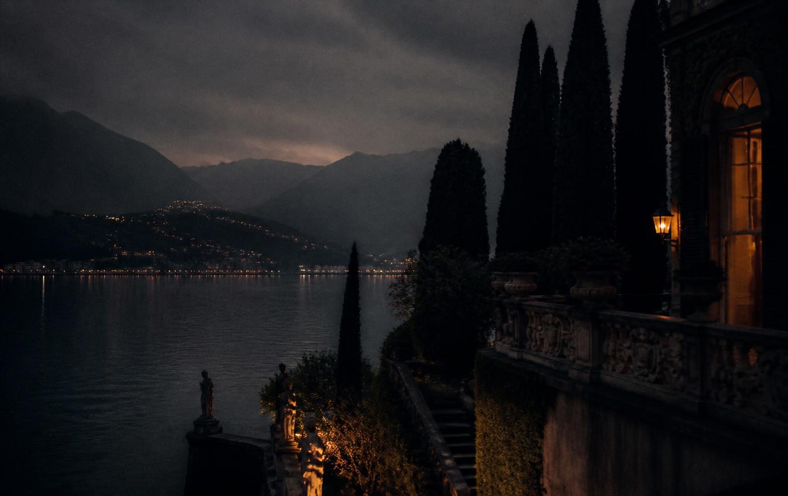 A cypress-lined villa terrace above Lake Como at dusk with lamplight and mist over the dark water