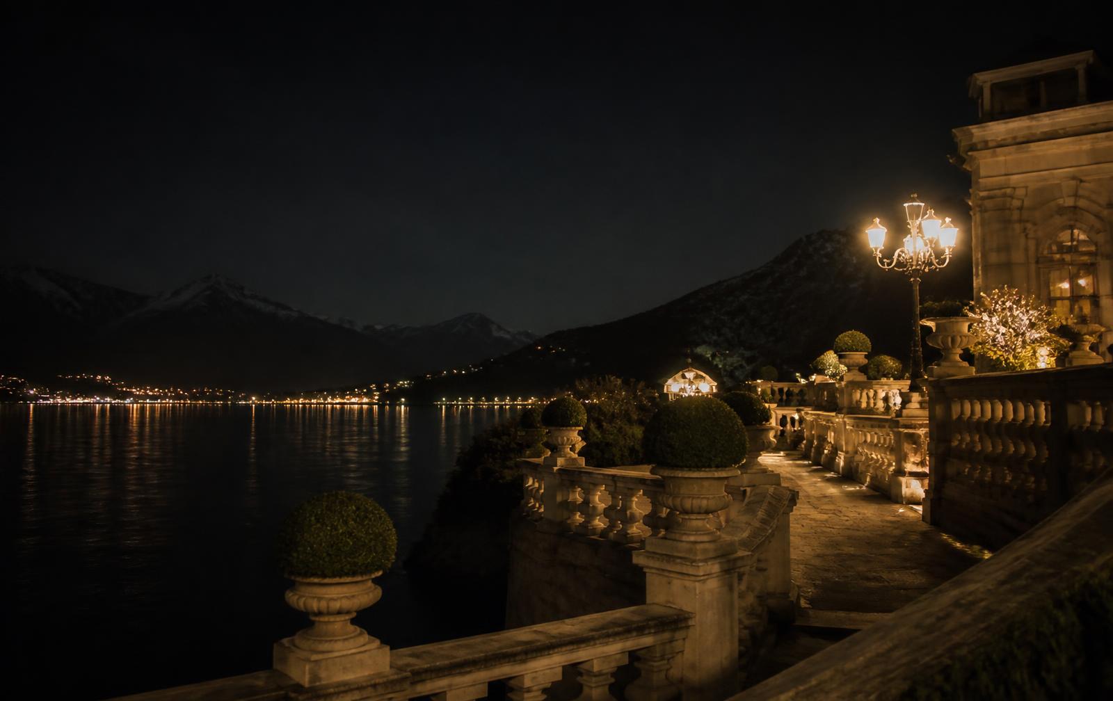 An elegant lakeside terrace at night above dark water with gold lamplight and distant alpine silhouettes