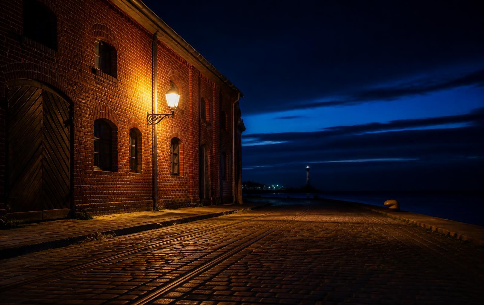 A red-brick harbour warehouse in Liepāja at night, a single lamp over cobbles, a distant lighthouse on a dark blue sea