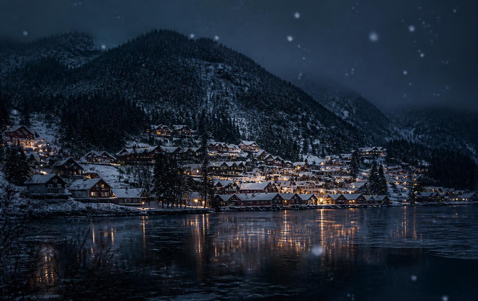 An inland Norwegian mountain town at night in falling snow, wooden buildings with warm lit windows above a dark lake, forested hills behind