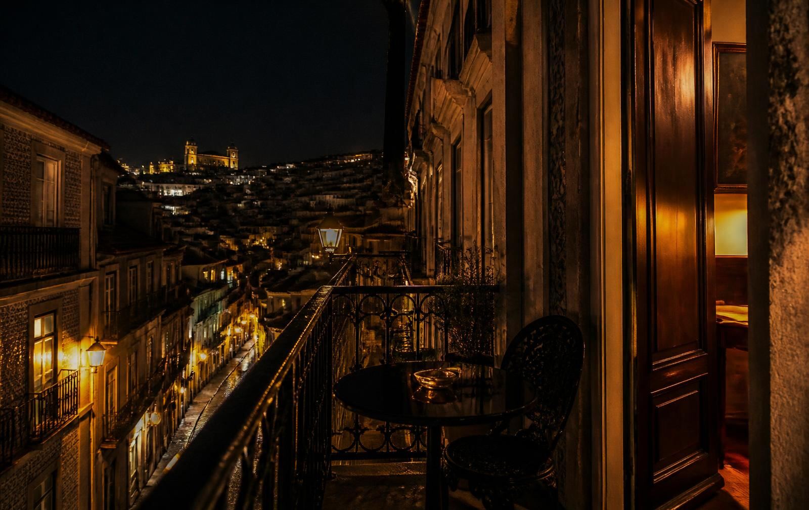 A wrought-iron balcony at night above lamplit Lisbon streets with warm interior light and dark wood behind