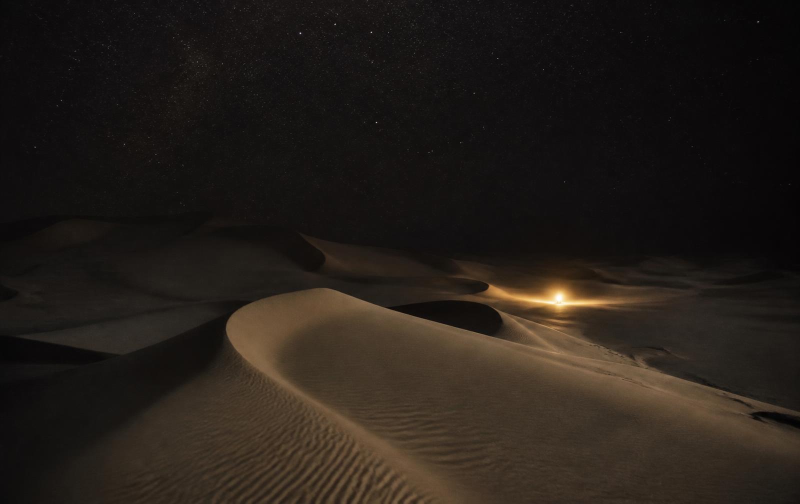 Vast empty desert dunes at night under a star-filled sky with a single distant warm gold light
