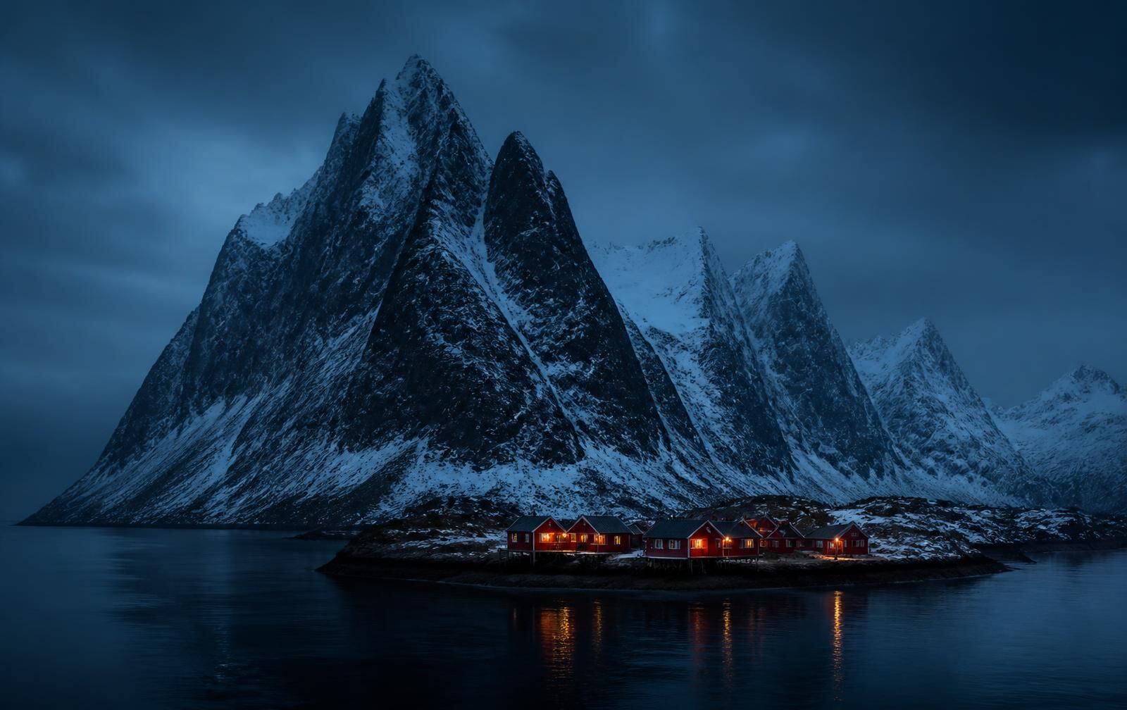 Jagged snow-covered Arctic peaks rising from a dark sea at blue hour, a small cluster of red wooden houses lit warmly on the shore