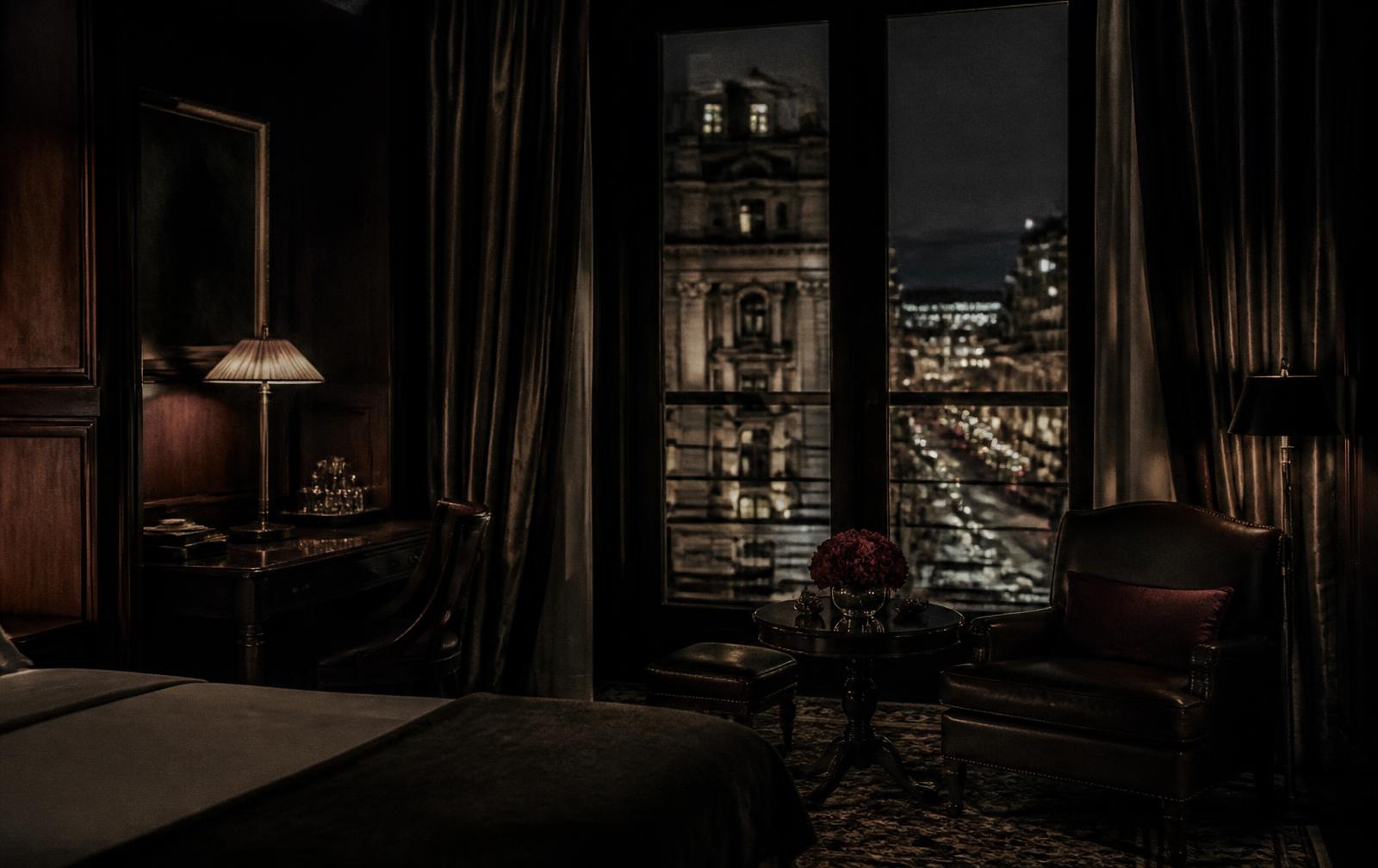 Dark panelled London hotel suite at night with a low lamp and city lights beyond tall windows