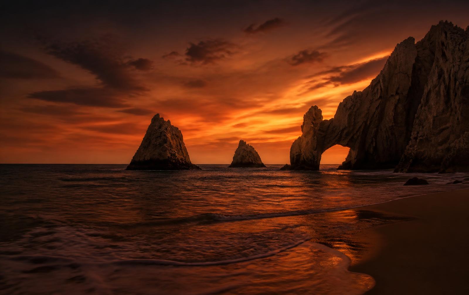 The rock arch of Los Cabos at dusk under an amber sky, gentle surf on the beach