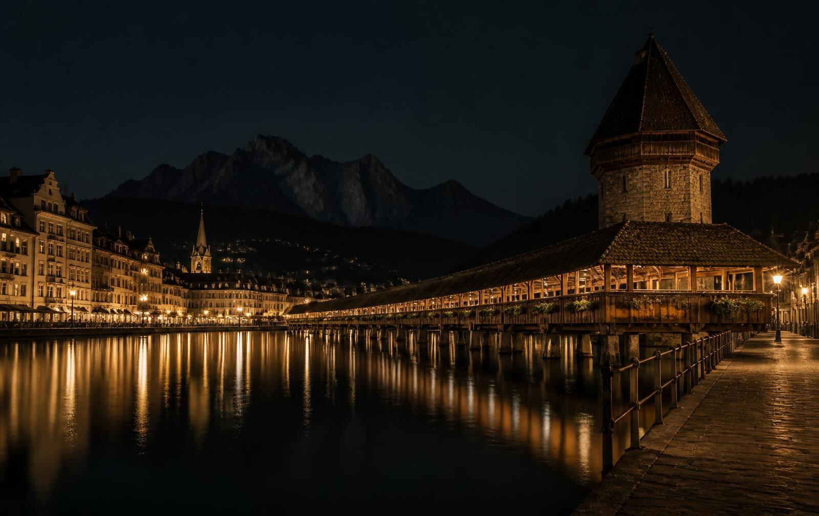 A historic lakeside old town at night with a covered wooden bridge and gold reflections on dark water