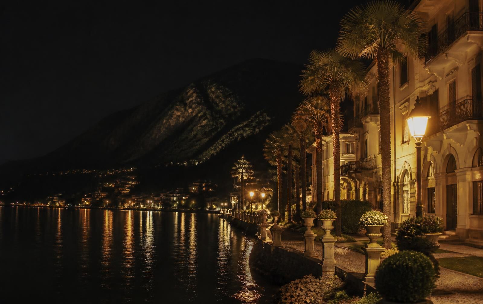 A warm southern lakeside promenade at night with palms, gold lamplight and a dark wooded mountain