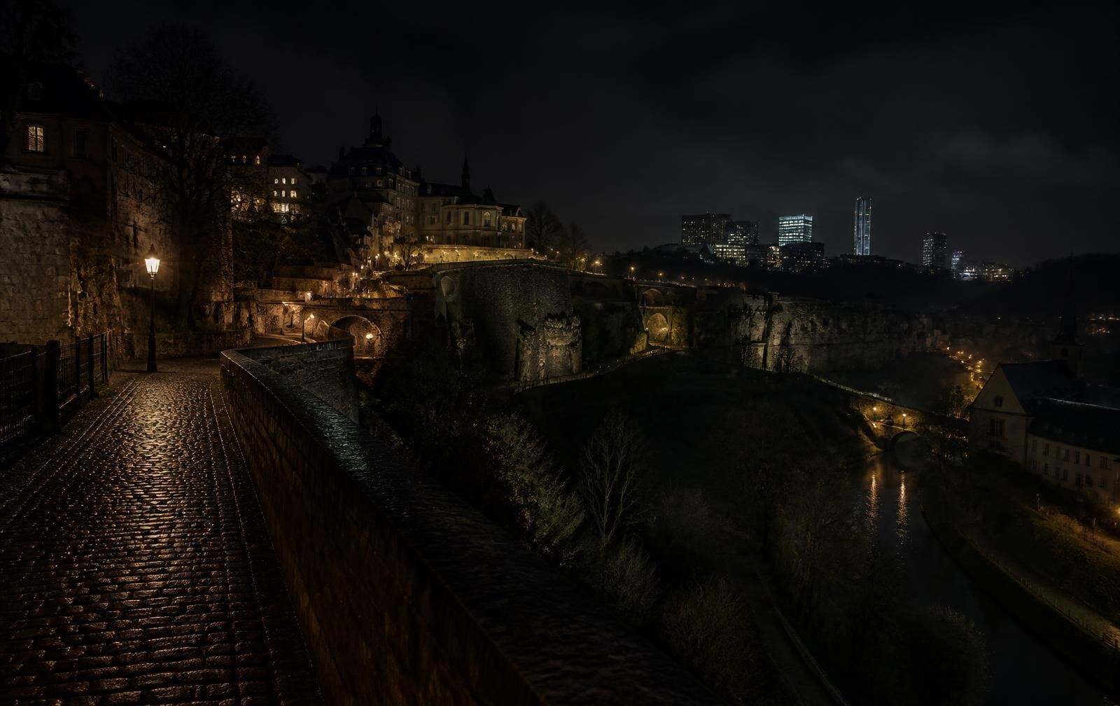 Old fortress walls above a dark river valley at night with modern glass towers lit in the distance