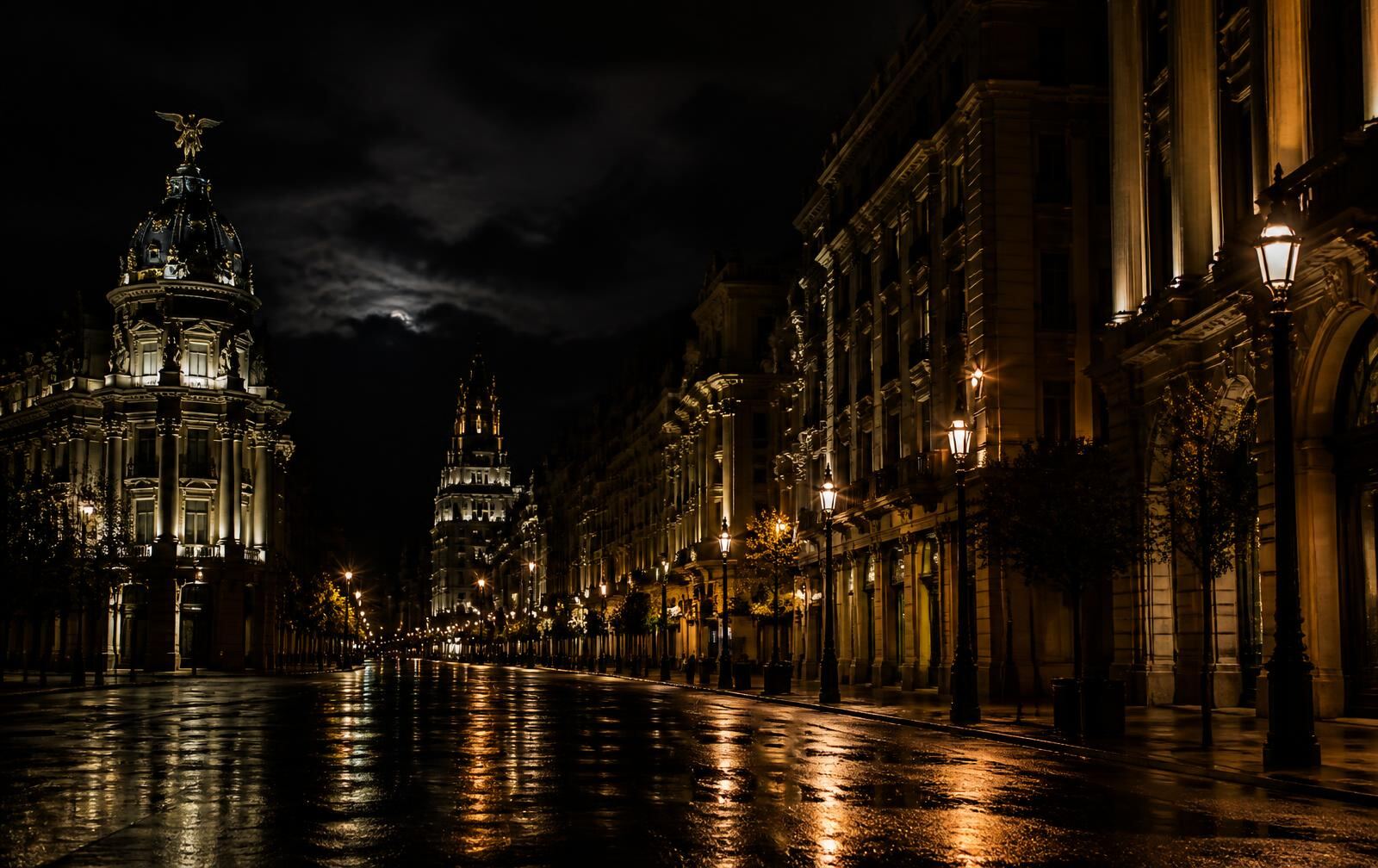 A grand Madrid boulevard at midnight with warm golden streetlamps on wet stone and lit facades