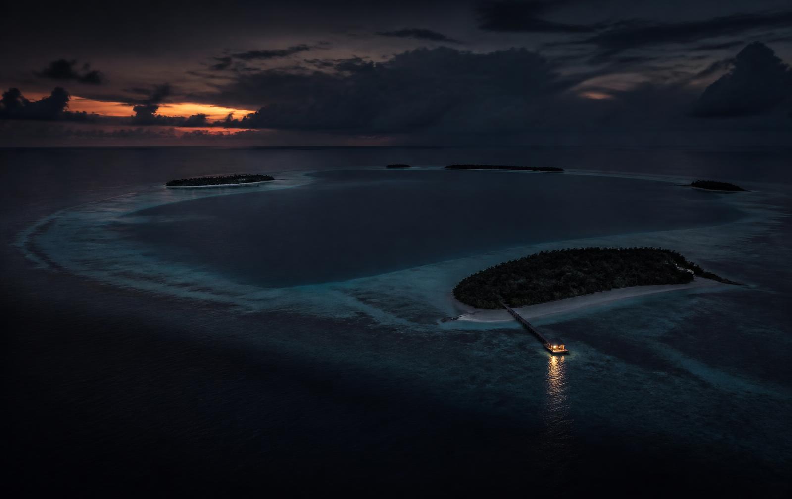 An aerial view of a Maldivian atoll at late dusk with dark indigo ocean and one lit jetty