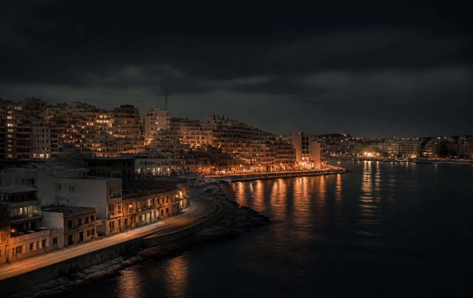 A dense low-rise island city at night, warm amber street lights reflecting across dark harbour water