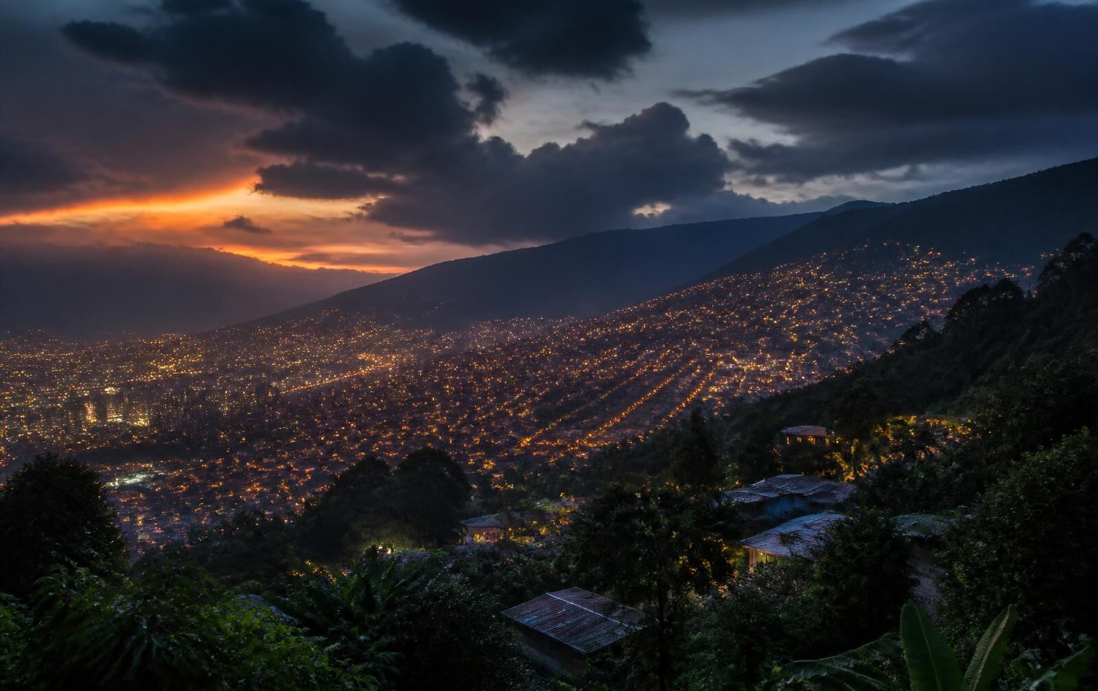 Medellín at dusk, warm city lights spread across green mountain slopes under dramatic evening clouds