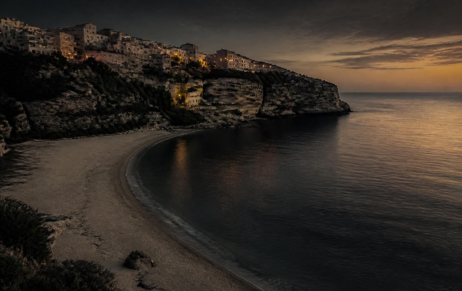 A northern Mediterranean coastline at dusk with a sandy bay below a limestone hillside town showing scattered warm lights