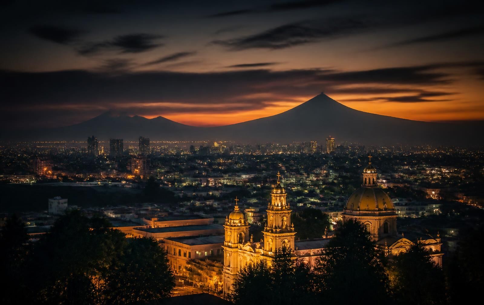 Mexico City skyline at dusk with a lit colonial church in the foreground and volcano silhouettes against an amber sky