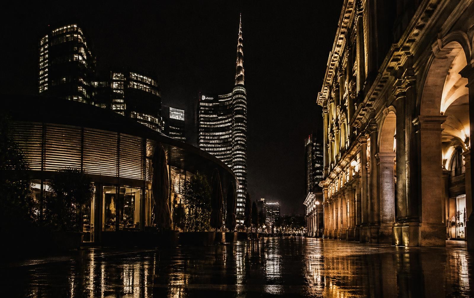 Rain-washed Milan streets at night with lamplit arcades and illuminated modern towers in the distance
