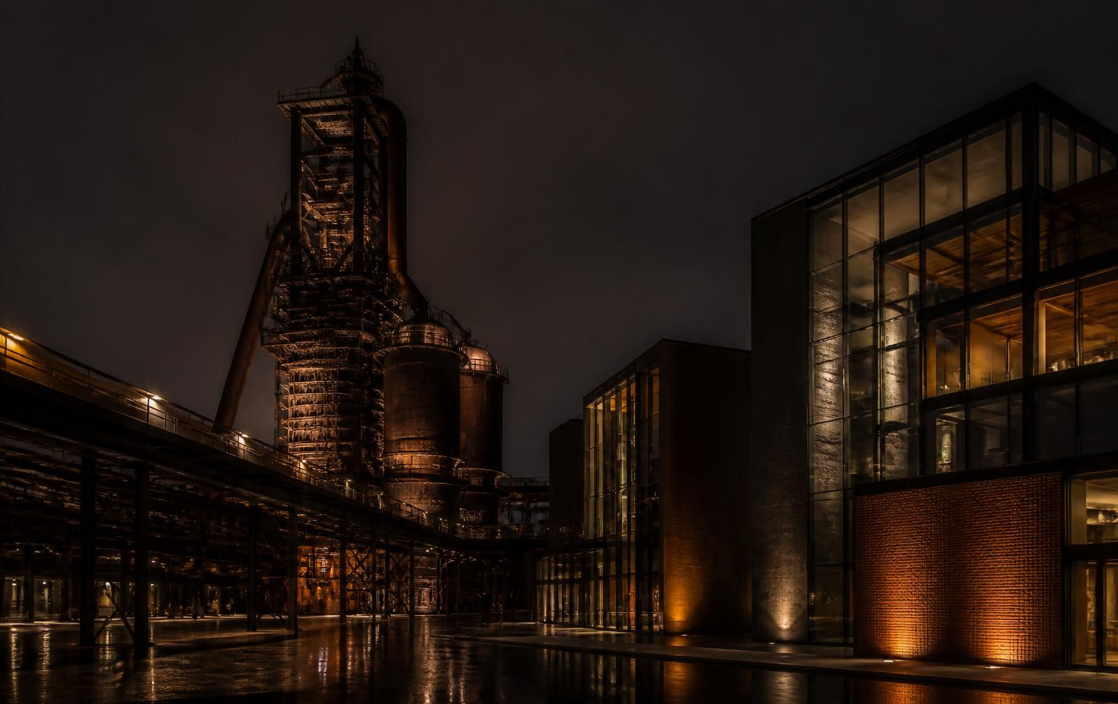 A former steelworks structure lit at night beside modern glass buildings with warm lit windows on a wet plaza