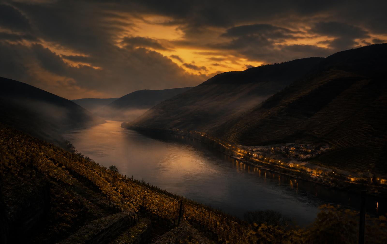 A wide river valley at dusk with terraced vineyards on the slopes and a few warm village lights near the water